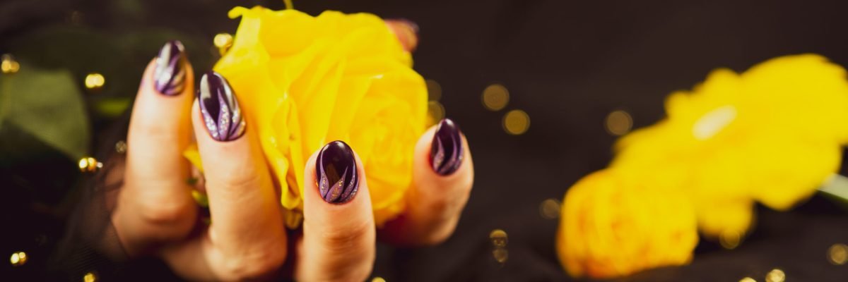 Adult woman's hand with purple manicure on black background. Crop unrecognizable person with design on nails holding yellow flower